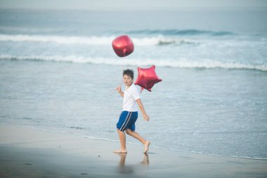 Mutlu çocuk plajda kırmızı balonlarla oynar ve yaz tatillerinde harika vakit geçirir. Yaşam tarzı, tatil, mutluluk, eğlence konsepti