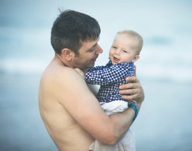 Neşeli genç baba ve küçük oğlu deniz kenarında birlikte eğleniyorlar, baba okyanusta çocuğu öpüyor.