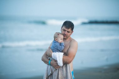 Neşeli genç baba ve küçük oğlu deniz kenarında birlikte eğleniyorlar, baba okyanusta çocuğu öpüyor.