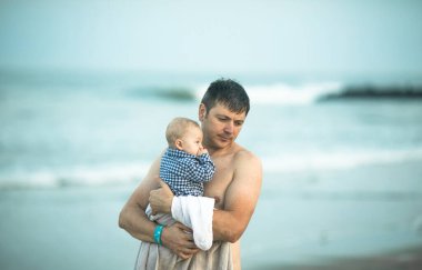 Neşeli genç baba ve küçük oğlu deniz kenarında birlikte eğleniyorlar, baba okyanusta çocuğu öpüyor.