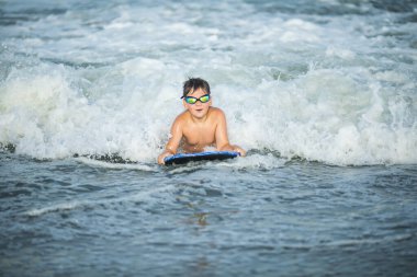 Küçük sörfçü deniz dalgasında sörf yapmayı öğreniyor. Çocuk yaz okyanusunda oynuyor, sörf öğreniyor, dalgalara biniyor. Küçük çocuk sörf tahtasında yüzüyor.