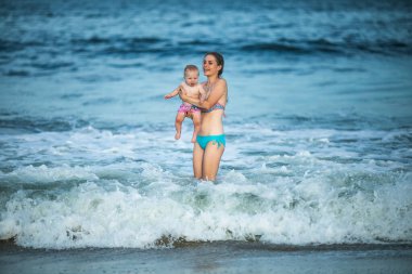 Anne, okyanus dalgasında bebeğine sarılıyor. Genç bir anne ve güzel çocuğu arasındaki bağ ve birliktelik kavramı..