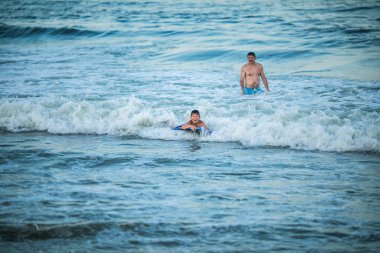 Küçük sörfçü deniz dalgasında sörf yapmayı öğreniyor. Baba ve oğul yaz okyanusunda oynuyorlar, sörf öğreniyorlar. Küçük çocuk sörf tahtasında yüzüyor.