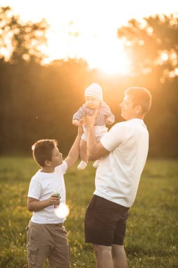 Mutlu aile kucaklaşmaları ve yaz yürüyüşlerinde kucaklaşmaları. Baba ve iki oğlu parkta yürüyor ve güzel doğanın tadını çıkarıyorlar. Sevimli bir aile fotoğrafı..