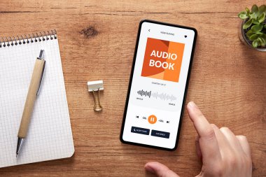 Akıllı telefonlu bir el resmi, rahat çalışma sahasında sesli kitap çalıyor. Ses kitabı uygulama kavramı.