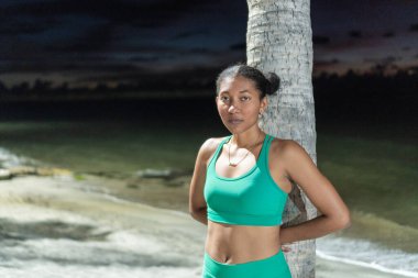 Spor kıyafetli esmer bir kadın ağaca yaslanmış ve kameraya bakıyor..