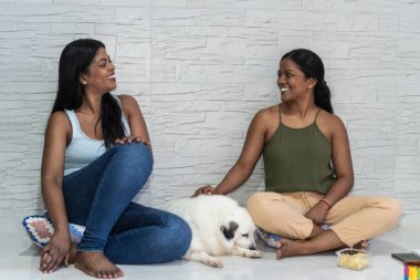 İki genç kadın köpekleriyle birlikte evlerinin zemininde oturuyorlar.