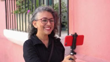 Latin female influencer smiling while broadcasting with her cell phone outdoors on the street.