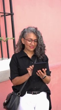 Hispanic woman in glasses holds tablet outdoors.