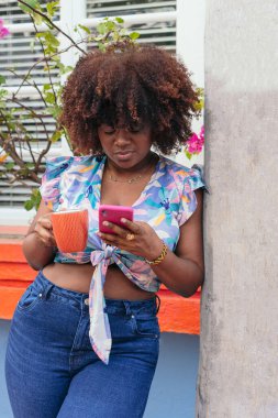 Afro kadın kahveden zevk alıyor ve sokakta cep telefonuna bakıyor..