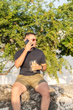 Elinde atıştırmalık bir şeyler tutarken, arka planda ağaçlarla dışarıda oturan sıradan bir adam..