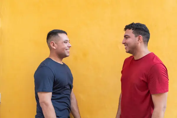 Two men in casual attire, one in a dark shirt and the other in red, enjoy a light-hearted conversation with a bright yellow wall behind them.