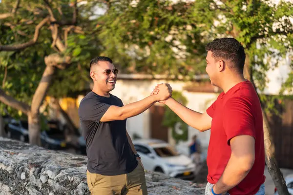 Two smiling men greet each other with a handshake outdoors, radiating friendship.