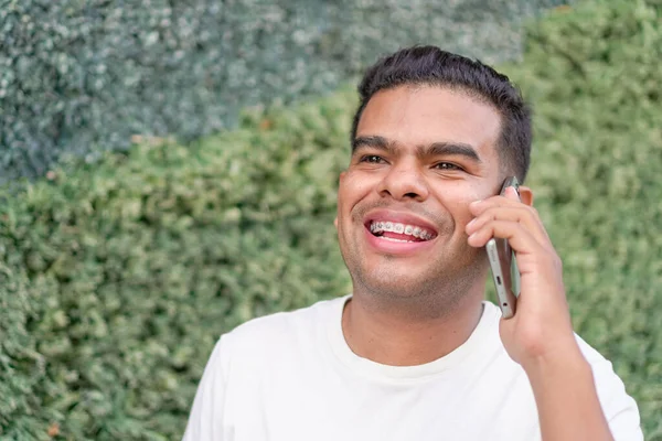 Smiling young man with dental braces enjoying a conversation on his smartphone, outdoor with a green hedge in the background.