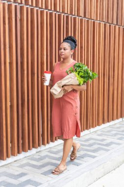 Elinde kahve fincanı ve taze yeşillikler olan bir kadın şehirde yürüyor..