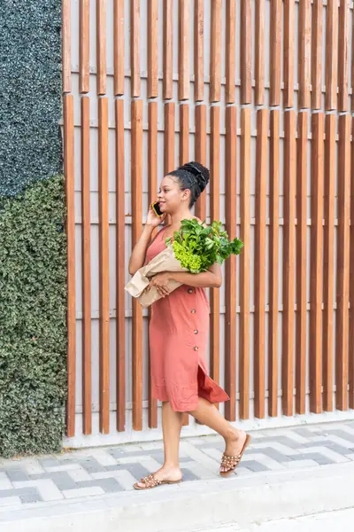 Multitasking woman chatting on a mobile phone while carrying leafy greens.