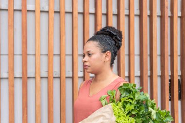 Bir çanta dolusu yapraklı yeşillikle modern ahşap bir çite karşı düşünceli bir kadın..