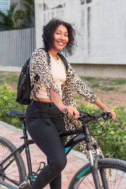 Kıvırcık saçlı neşeli genç bir kadın bisikletinin yanında duruyor, leopar desenli ceket ve sırt çantası giyiyor..