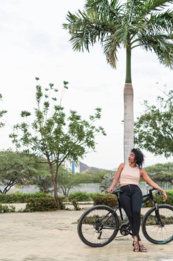 Rahat bir kadın bisikletiyle, bereketli tropik yeşilliklerin ve sakin bir parktaki palmiye ağaçlarının arasında duruyor..