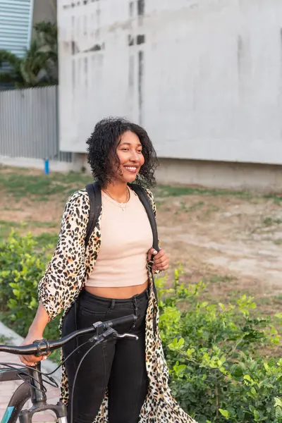 Kıvırcık saçlı Latin kadın bisikletinin yanında duruyor, leopar desenli ceket ve sırt çantası giyiyor..