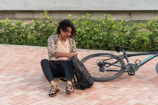 Düşünceli genç bir kadın bisikletinin yanındaki tuğla yolda oturur ve sırt çantasından bakar..