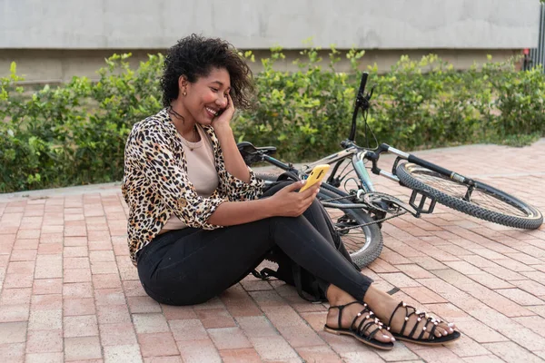 Gülümseyen bir kadın yerde oturuyor, telefonundaki mesajın tadını çıkarıyor, yanında bisikletiyle..