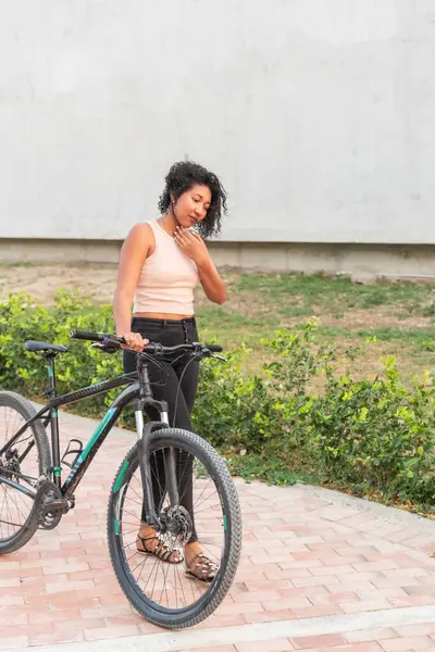 Düşünceli bir kadın bisikletiyle duruyor, arkasında yeşillik olan asfalt bir yolda düşüncelere dalmış..