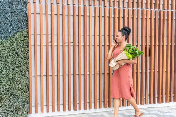 Woman multitasking with a phone call and fresh groceries in hand against an urban backdrop.
