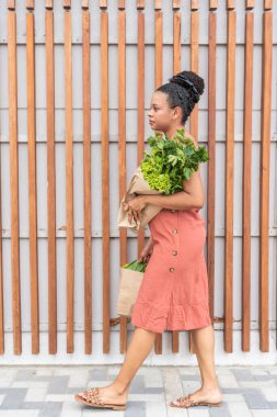 Somon elbiseli genç bir kadın kaldırımda taze yeşillikler taşıyor.