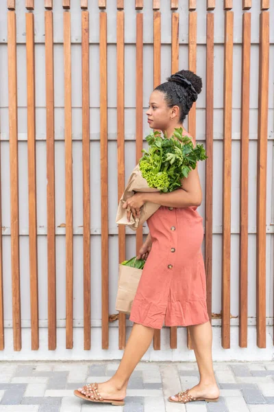 Somon elbiseli genç bir kadın kaldırımda taze yeşillikler taşıyor.