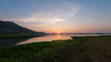 Sakin bir göle yansıyan nefes kesici bir gün batımı. Dağların siluetleri ve ön planda yemyeşil bitkiler..