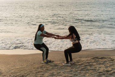 Spor kıyafetli Latin kadınlar kumlu bir sahilde çömelirken el ele tutuşuyorlar.