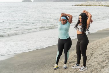 Okyanus manzaralı sahilde kol esnetme egzersizleri yapan iki kadın..