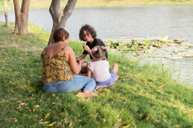 Göl kenarındaki çimlerin üzerinde oturan aile, açık havanın tadını çıkarıyor..