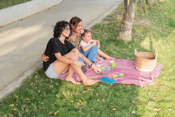 Bir anne ve çocukları piknik battaniyesinde gülüp sarılıyorlar, parkta güzel bir günün tadını çıkarıyorlar..