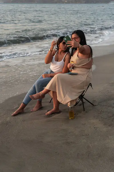 stock image Two women posing for a playful selfie while toasting with wine on a sandy beach at twilight.