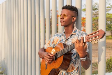 Şık güneş gözlüklü ve desenli gömlekli düşünceli Afrikalı genç adam dışarıda gitar çalıyor, arka planında modern metal bir çit ve günbatımı ışığı var..