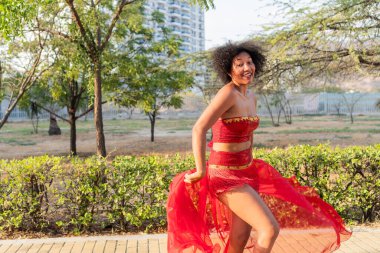 Canlı kırmızı elbiseli bir kadın, yemyeşil ve modern gökdelenlerle çevrili, neşeli ve kaygısız bir anı yakalayan şehir parkında neşeyle dans ediyor..