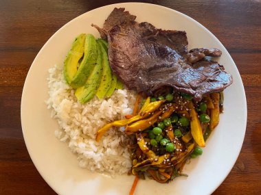 Beyaz bir tabakta servis edilen ızgara sığır eti, beyaz pirinç, avokado dilimleri ve bezelyeli kızarmış sebzelerin yer aldığı tabağın genel manzarası..