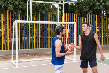 Açık bir sahada renkli çitlerle çevrili bir sahada konuşan ve basketbol oynayan iki adam..