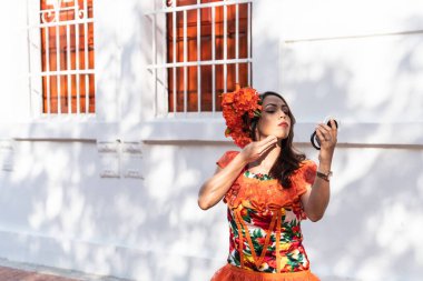 Canlı, çiçek desenli turuncu bir elbise giymiş bir kadın ve aynayla uyumlu bir başlık takıyor. Parmaklıklı pencereli beyaz bir duvarın önünde duruyor..