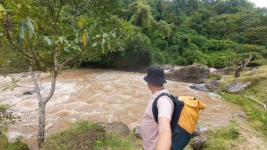 Şapka ve sırt çantası takan bir yürüyüşçü sık, yeşil bir ormanda hızla akan bir nehre hayran kalır. Açık hava macerasını ve doğa takdirini temsil eder..