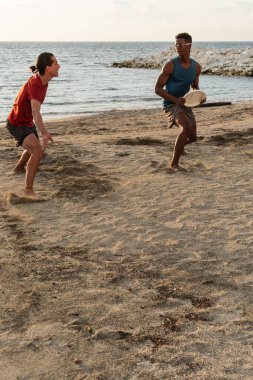 Gün batımında okyanusun kıyısında kumlu bir sahilde frizbi oynayan iki adam..