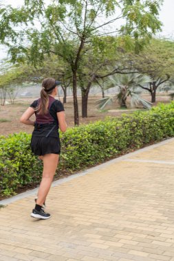 Ağaçlar ve yeşilliklerle çevrili bir parkta kaldırımlı bir yolda koşan bir kadın..