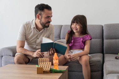 Baba ve kızı kanepede bir hikaye paylaşıyor, birlikte sıcak bir okuma anının tadını çıkarıyorlar..