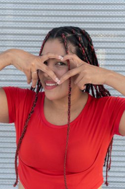 Metal bir duvarın önünde elleri ve parmaklarıyla kalbi şekillendiren gülümseyen genç kadın.