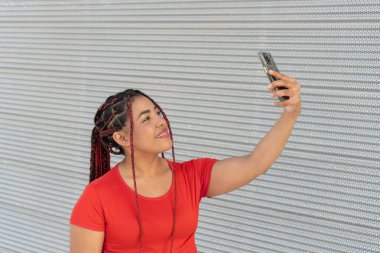 Genç Latin kadın, metal duvarın önünde akıllı telefonuyla selfie çekiyor.
