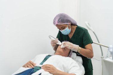 Güzellik uzmanı güzellik kliniğinde sedyede yatan bir hastaya yüz bakımı için bir cihaz kullanıyor.