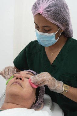 Cerrahi maske takan ve eldiven takan bir güzellik uzmanı güzellik salonunda hastanın yüzüne makyaj çıkarıcı pedler takıyor.