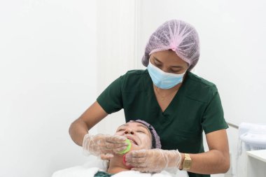 Güzellik uzmanı ameliyat maskesi ve eldiven takıyor. Güzellik kliniğinde yüz bakımı sırasında hastanın yüzüne kozmetik ürünler uyguluyor.